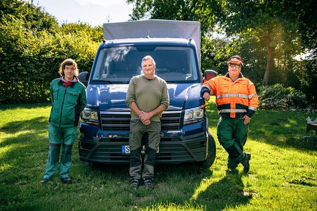 Drei Leute in Garten-Montur vor einem Transporter lachen.