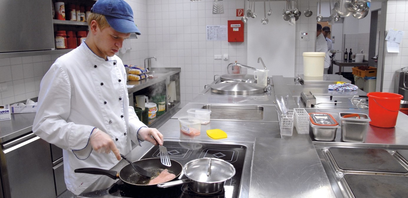 Das Bild zeigt einen jungen Mann, der an einem Herd steht und Essen in einer Pfanne zubereitet.